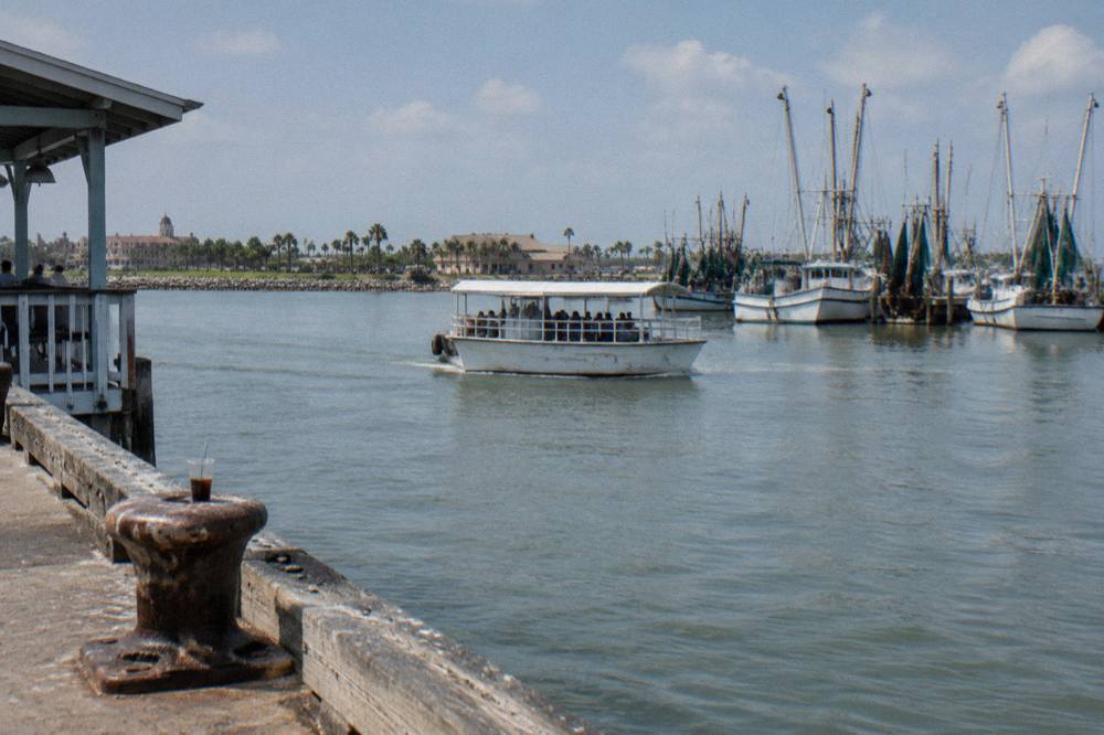 A covered passenger boat gliding across a calm Galveston harbor channel near docked shrimp boats