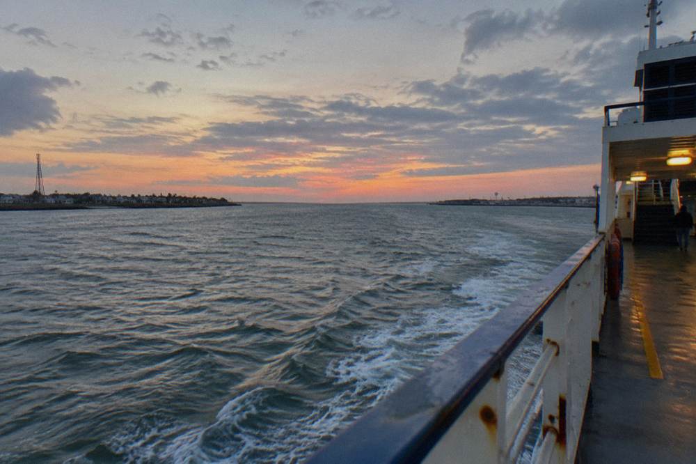 A ferry crossing open channel water between two shorelines under a wide evening sky