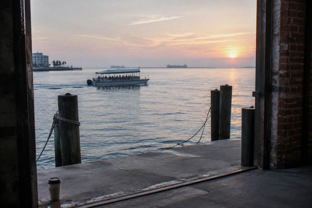 A covered tour boat crossing calm Galveston Harbor near piers with mooring lines and distant ships