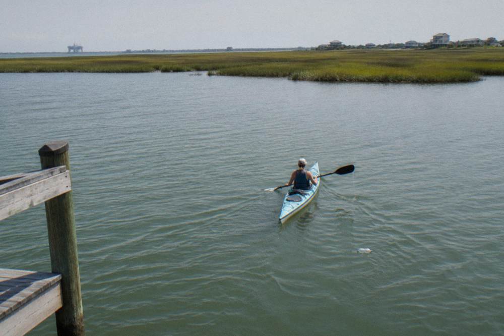 A small kayak gliding across calm green bay water toward a distant shoreline of marsh grass