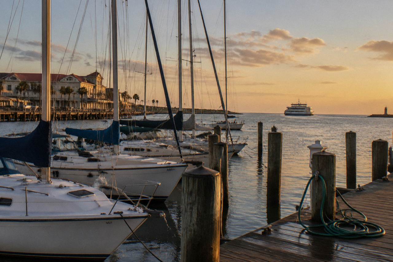 A calm Galveston harbor at golden hour with moored sailboats and a distant ferry crossing the channel