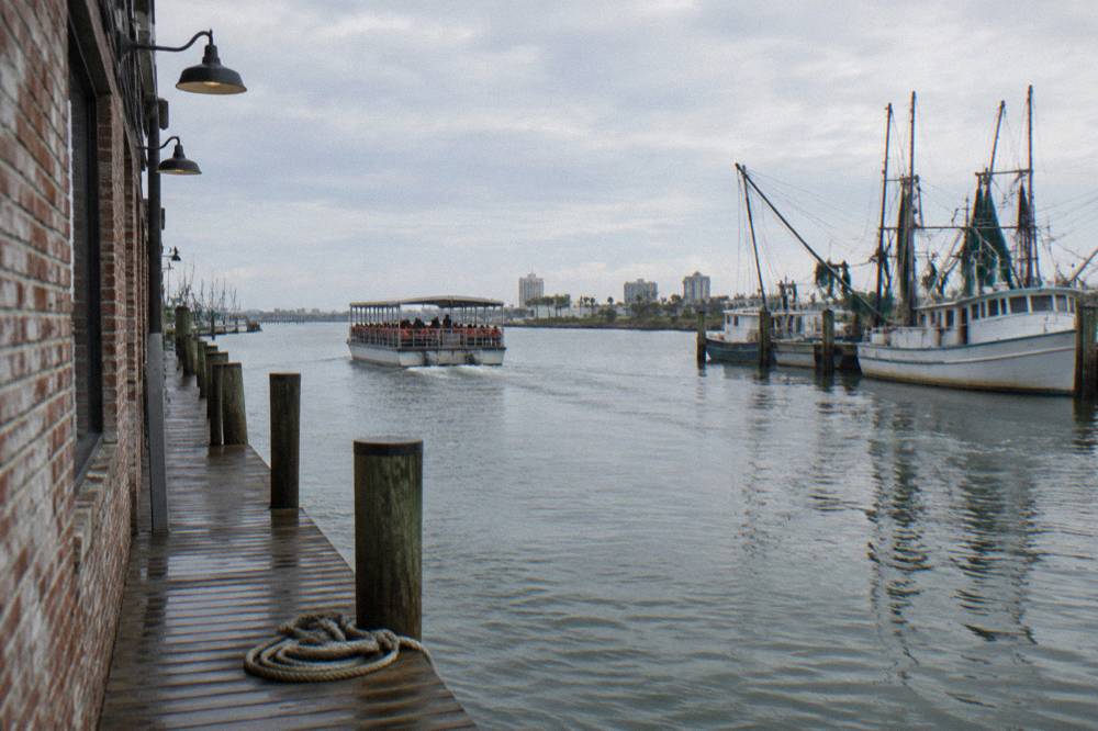 A covered tour boat with passengers at the rail moving through a harbor channel past moored fishing vessels