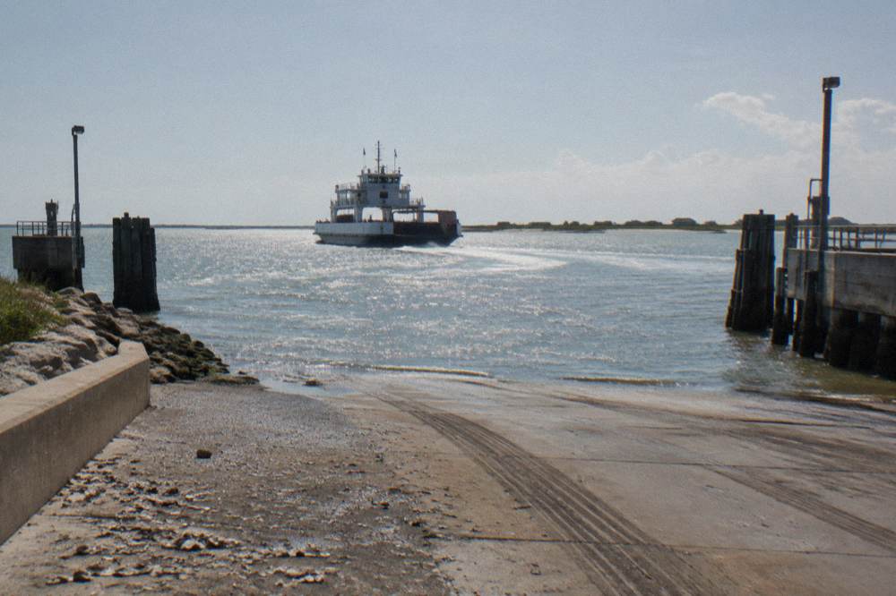 A ferry boat crossing a wide ship channel with a distant shoreline and open sky