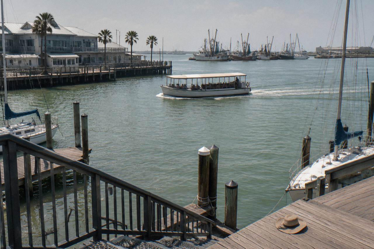 A small covered tour boat crossing calm Galveston harbor water near a wooden pier