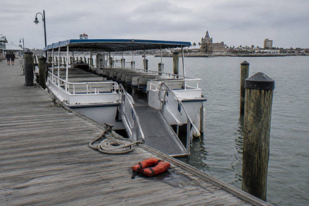 A covered excursion boat docked at a Galveston pier, boarding ramp down, harbor water behind it
