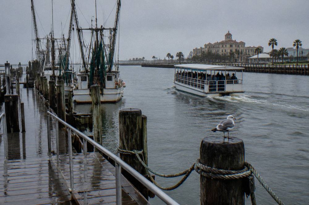 A covered tour boat moving through Galveston harbor past docked ships