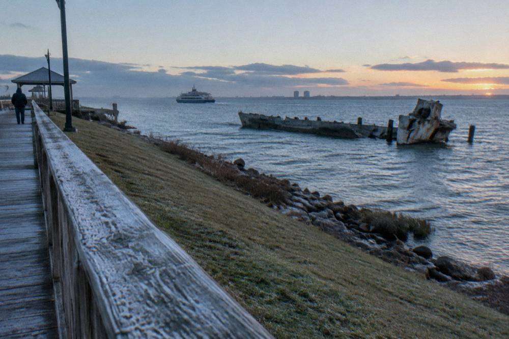 A grassy shoreline park overlooking a ship channel with a pier and passing vessels