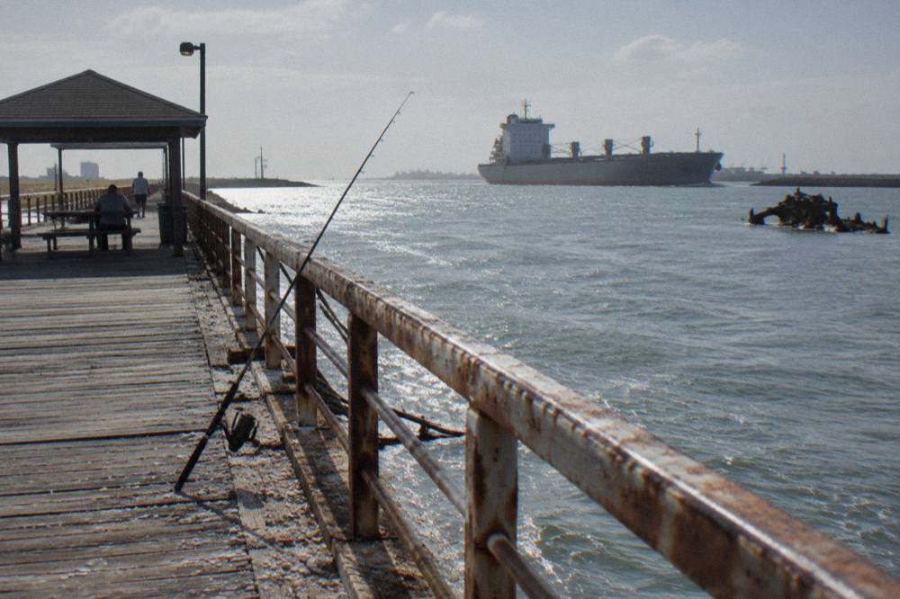 A fishing pier over calm channel water with a large ship passing in the distance
