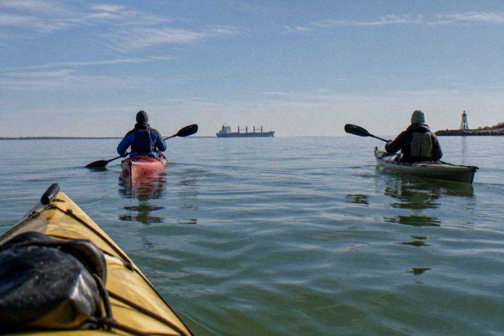 Two kayakers paddling across a wide calm bay near a distant ship channel