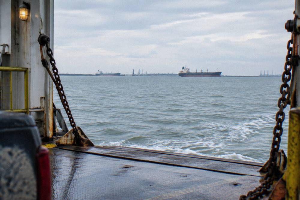 A vehicle ferry underway on open channel water with distant shoreline and moored ships