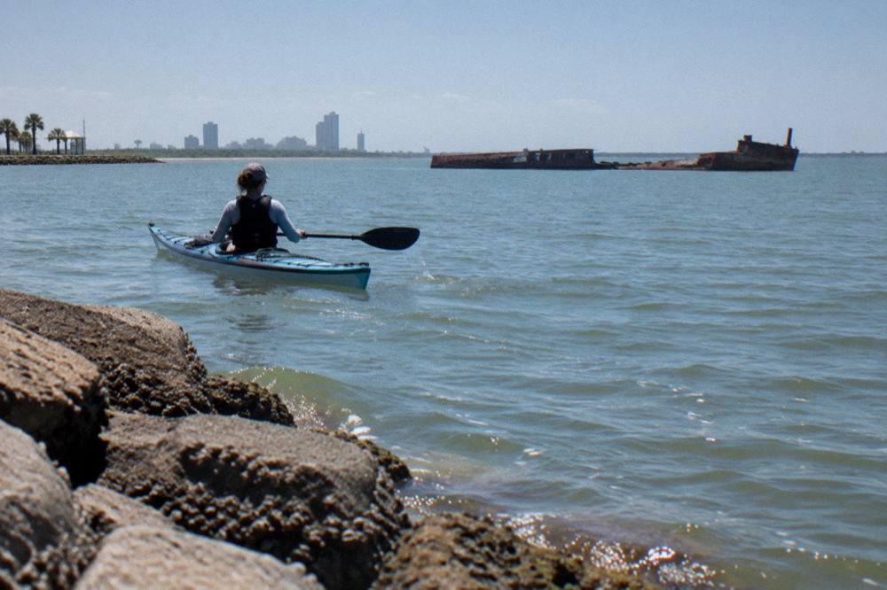 A sea kayak gliding across calm harbor water near a Galveston shoreline at midday