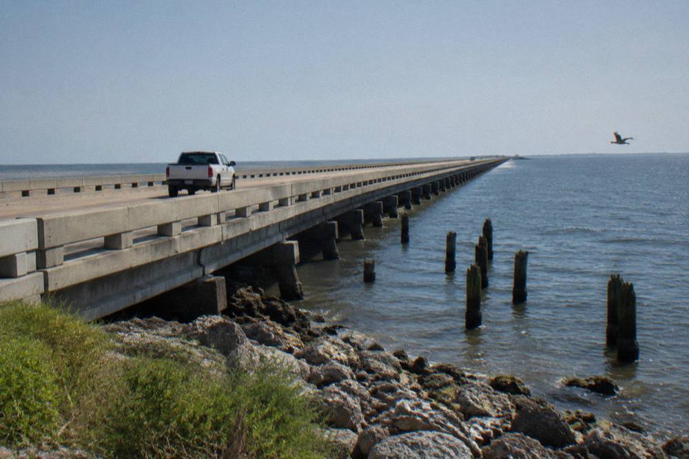 A pickup truck crossing a long causeway bridge toward a barrier island with open water on both sides