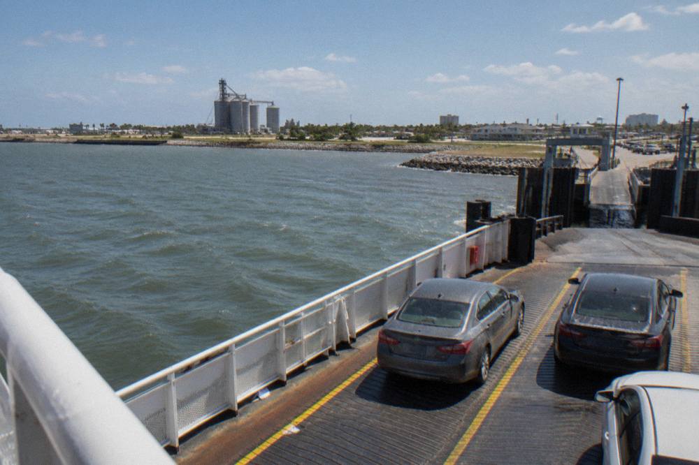 Cars lined up on a ferry deck with the Galveston shoreline behind them under a wide sky