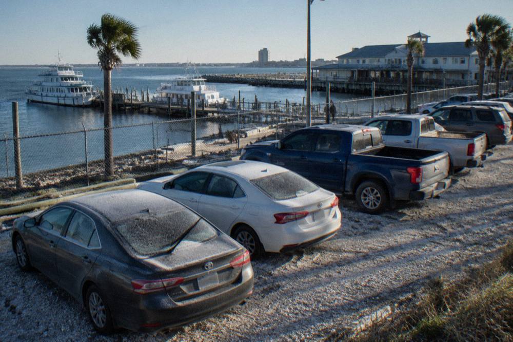 Cars parked in a waterfront lot beside a harbor road with tour boats at a nearby pier