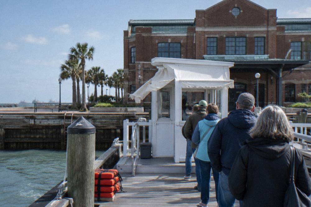 A white ticket kiosk beside a hotel entrance with a boarding line of visitors at a harbor pier