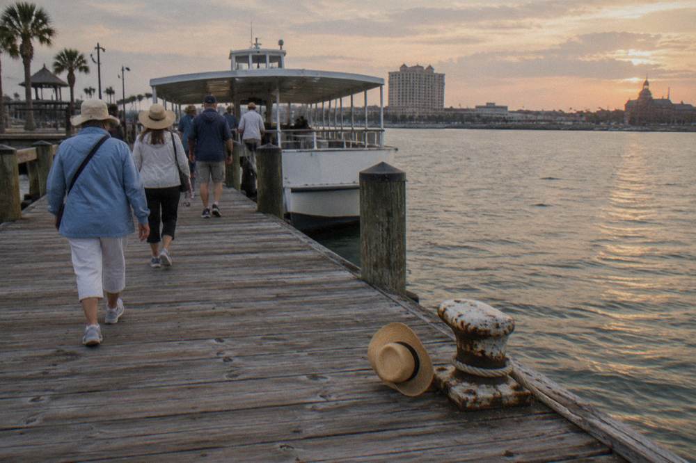 Visitors in sun hats and light jackets walking down a wooden pier toward a covered tour boat