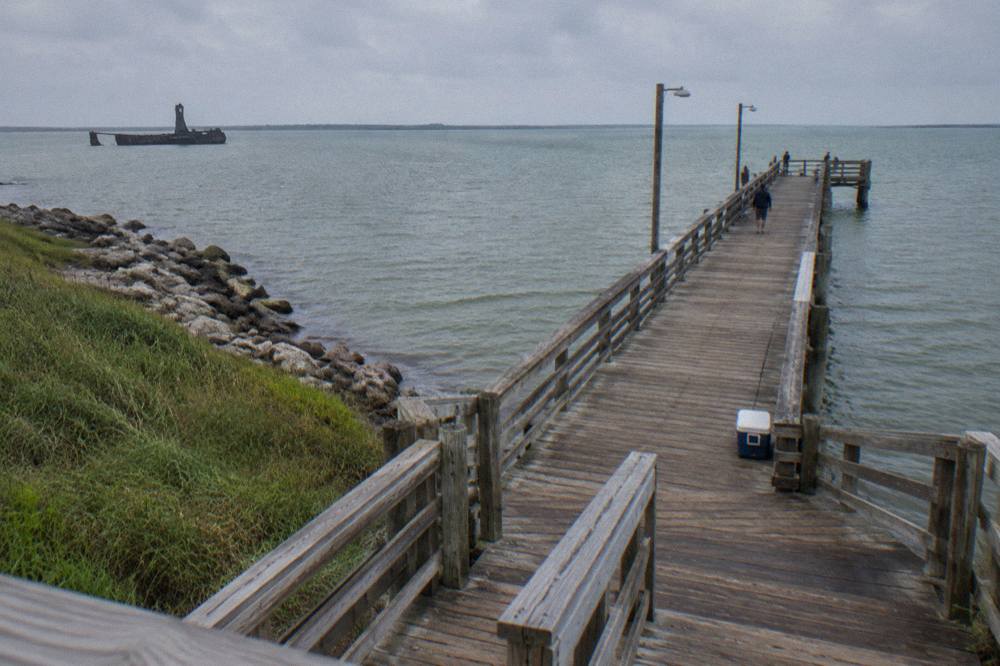 A long wooden fishing pier extending over calm green harbor water under a wide sky