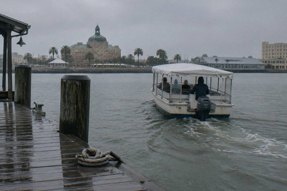 A small covered tour boat pulling away from a wooden pier along a working harbor